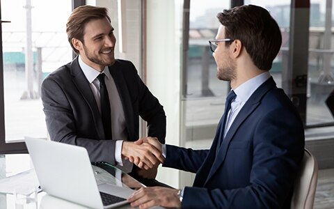 Two male professionals shaking hands, smiling, and making a deal at an office.