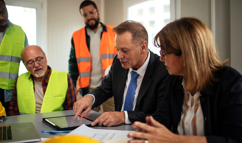 a group of people who work in the construction industry having a meeting, looking at plans and a digital table on a desk