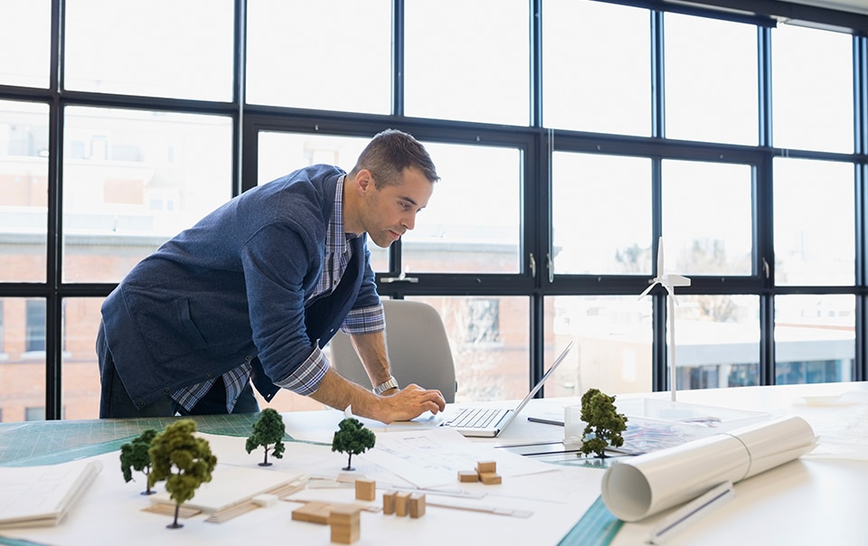 man standing at his desk while working on laptop, how to choose a professional liability insurance policy for your design firm