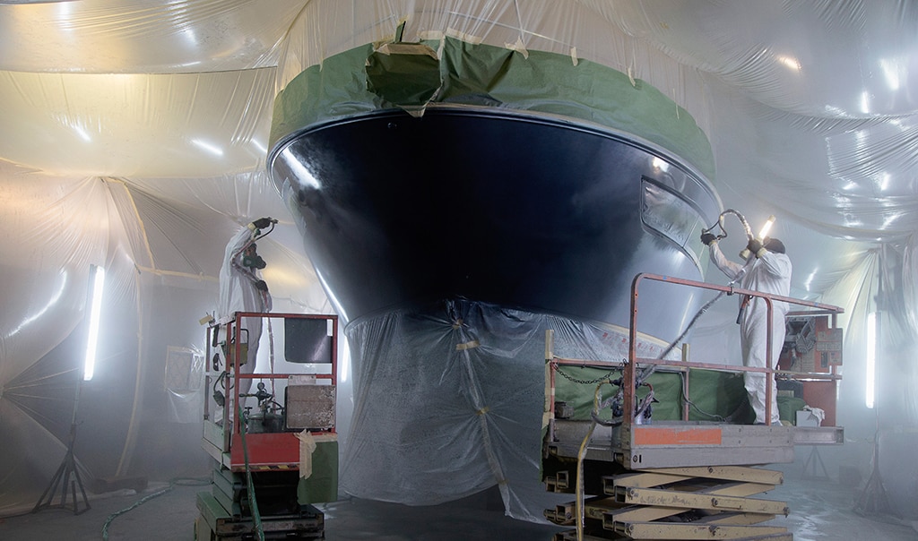 Two men paint boat in warehouse.