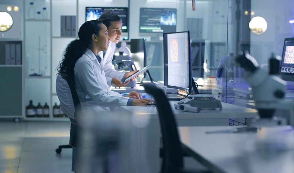 Two women working on a large desktop and tablet in a science lab.