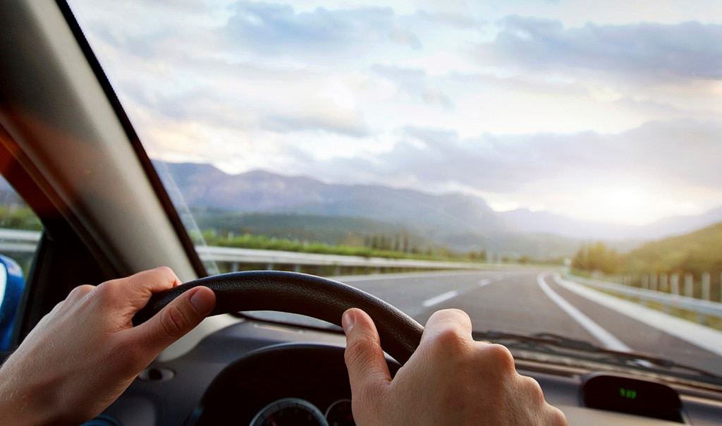 hands on the steering wheel of a car, a road and mountains in the background