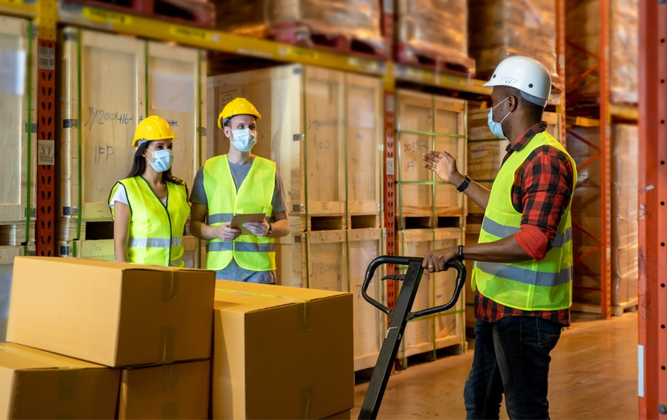 Three workers in a warehouse talking to each other with safety vests, hard hats and face masks on. 6 Evolving Risks Impacting Workplace Injuries