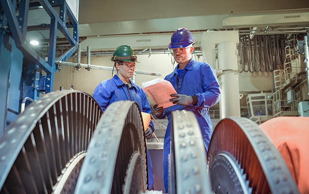 Two workers in safety gears having a discussion and checking a machine with gears.