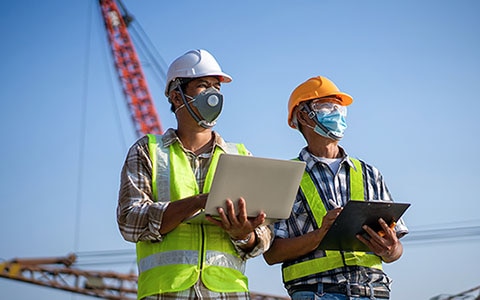 Two workers at a construction site holding laptop computers, wearing safety equipment