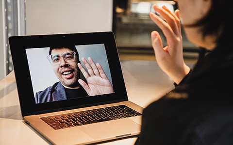 Two people sitting at their desk waving at each other during a virtual meeting.