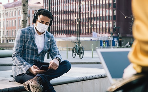 man sitting on bench in city with a mask on and looking at his phone, 