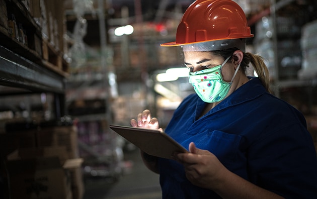 woman wearing hardhat and mask in warehouse while she is on tablet. Navigating Your Business Through COVID-19