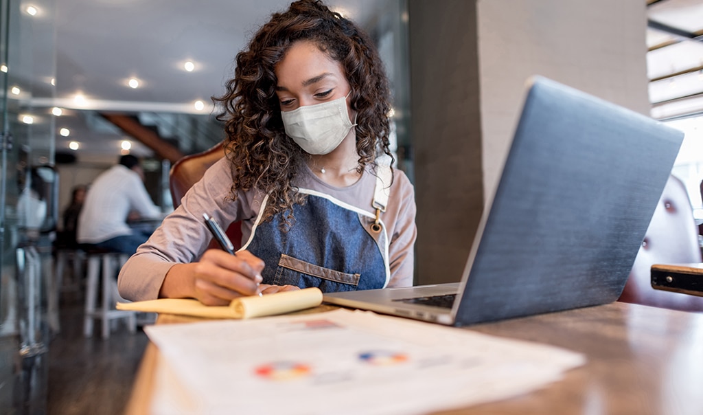 woman sitting at table in cafe with laptop and financial paperwork in front of her as she writes on a notepad. How to Protect Your New Business