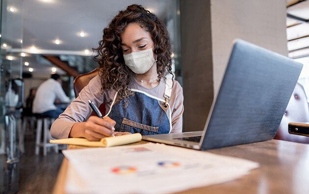 woman in a mask sitting at a table at a cafe with laptop in front of her as she writes on a notepad, How to Protect Your New Business