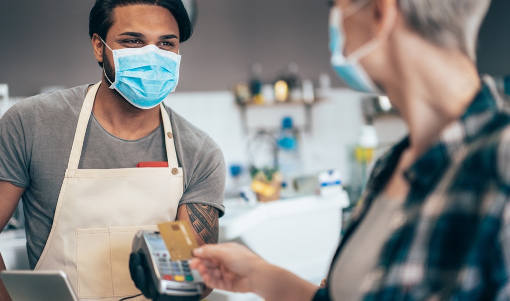 man in mask and apron holding a card reader up for a customer to use. The customer is also in a face mask. How to Identify Future Customers and Reward Top Current Customers