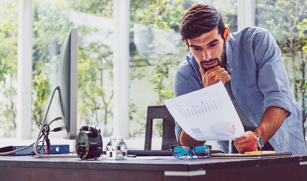 Man standing over desk with his elbow on his desk and his chin in his hand. He is looking at financial paperwork. Should You Pivot Your Business Model?