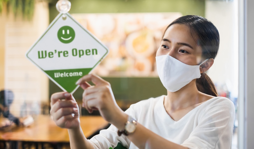 woman in a mask puts an open sign on her business door. How to Communicate Business Reopening with Customers