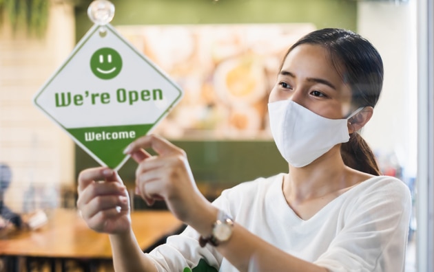 woman hanging open sign in doorway as she wears a mask. How to Communicate Business Reopening with Customers
