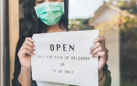 Woman wearing a mask puts a sign on the business door that says the business is only open for to go and pick up orders only. Small Business Compliance and Regulations Explained