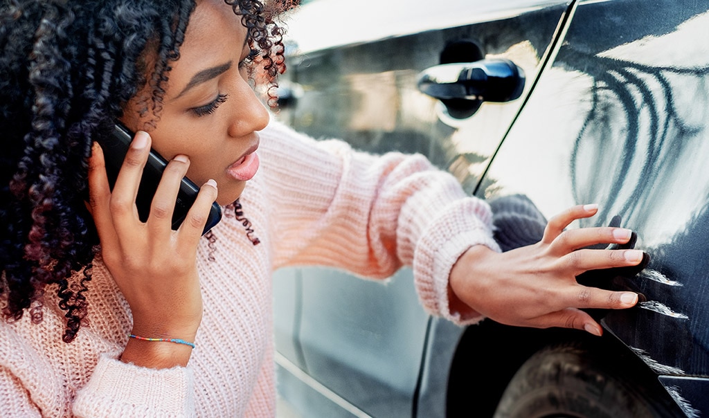 Young woman examining scratches and dents on her car, calling for help on her mobile phone