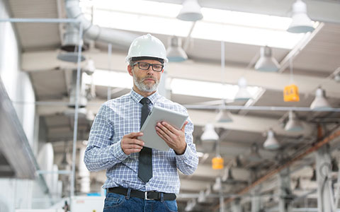Man in white hard hat and plaid shirt holding a tablet while walking in a warehouse.