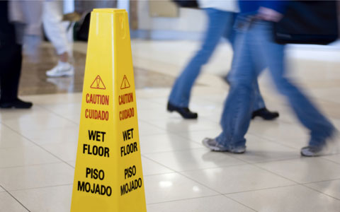 A yellow wet floor sign is posted on tile floor in an interior location that shows the legs of people walking.